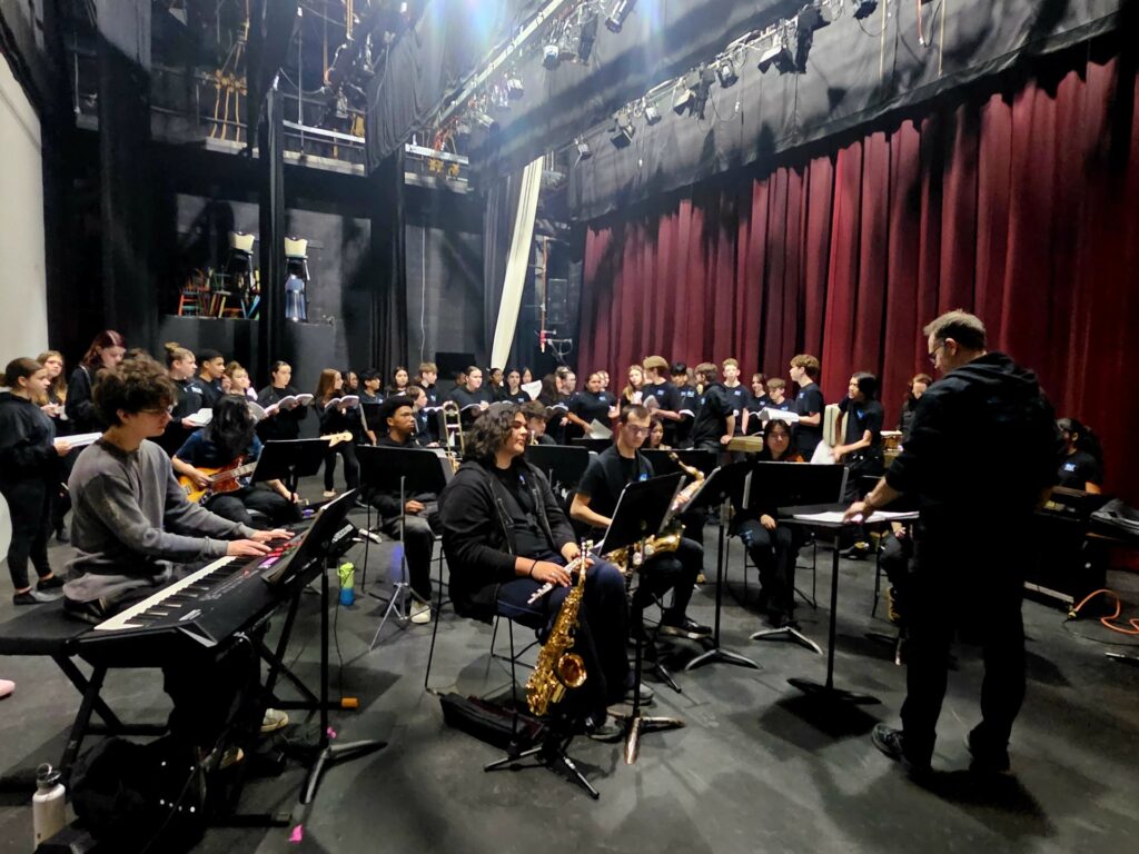 Oshawa Little Theatre Youth Group rehearsing Disney’s Newsies, with singers gathered behind a live student orchestra on stage.